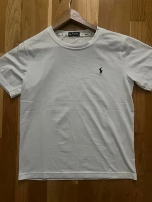 Vit t-shirt från Polo Ralph Lauren - Klassisk vit t-shirt från Polo Ralph Lauren med rund halsringning och korta ärmar. Liten mörkblå logga broderad på bröstet. Tillverkad i mjuk bomull för en skön och enkel stil.