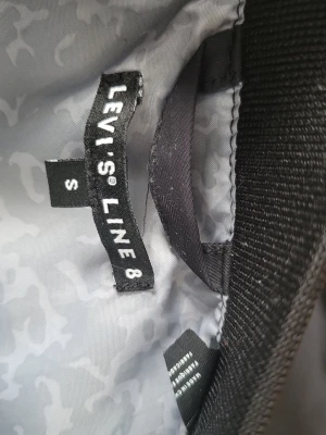 Svart bomberjacka Levi's Line 8 - Snygg svart bomberjacka från Levi's Line 8 i storlek S. Jackan har klassisk bombermodell med ribbade muddar och krage. Insidan har ett grått mönstrat foder som ger extra stilpoäng. Perfekt för dig som vill ha en clean och tidlös look. Använd 1 gång