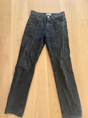 Svarta raka jeans från Zara - Säljer ett par svart/mörk gråa jeans från Zara med klassisk femficksmodell och raka ben. Jeansen har en enkel design med knappgylf och är tillverkade i ett slitstarkt denimtyg. Perfekta för dig som gillar en clean och tidlös look. Strl 36