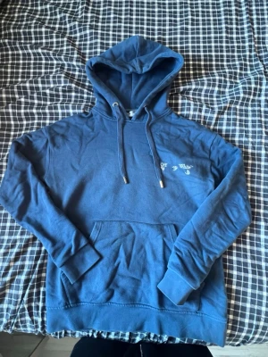 Blå hoodie från Off-White - Säljer en blå hoodie från Off-White med broderad logga på bröstet. Hoodien har justerbar dragsko i huvan och en stor magficka. Tillverkad i mjukt bomullsmaterial och har ribbade muddar vid ärmar och nederkant. Perfekt för en chill och stilren look.