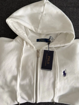 Vit hoodie från Polo Ralph Lauren - Klassisk vit hoodie från Polo Ralph Lauren med dragkedja framtill, huva med snörning och den ikoniska lilla loggan broderad i marinblått på bröstet. Tillverkad i mjuk bomull för maximal komfort. Perfekt för en clean och stilren look.
