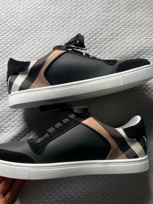 Burberry låga sneakers med rutmönster - Snygga låga sneakers från Burberry med klassiskt beige, svart och vitt rutmönster på sidan. Skorna har svarta läder- och mockadetaljer, vita sulor och svarta skosnören. Perfekta för dig som vill ha en stilren och exklusiv look.