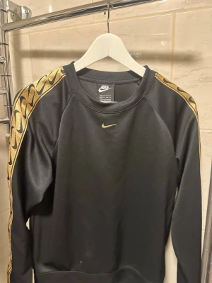 Svart Nike sweatshirt med guldmönster - Svart sweatshirt från Nike med guldfärgade detaljer längs ärmarna och liten gul Nike-logga på bröstet. Tröjan har rund halsringning och är tillverkad i ett mjukt material som passar perfekt till en sportig och trendig stil.