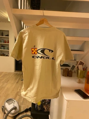 Beige O'Neill t-shirt med tryck - Beige t-shirt från O'Neill med stort logotryck på ryggen och mindre logga framtill. Klassisk passform med rund hals och korta ärmar. Perfekt för en avslappnad stil och enkel att matcha med jeans eller shorts.