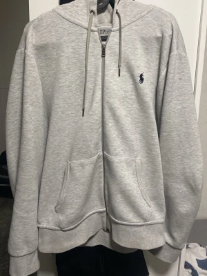Grå hoodie från Polo Ralph Lauren - Snygg ljusgrå hoodie från Polo Ralph Lauren med dragkedja, två stora fickor framtill och klassisk huva med snören. Ikoniska lilla loggan i mörkblått på bröstet. Tillverkad i mjukt bomullsmaterial som är perfekt för chill dagar. Inte använd så mycket, köpt på pondus för 1 år sen