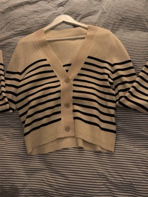 Beige och svart randig kofta - Snygg beige kofta med svarta horisontella ränder och v-ringning. Koftan har en croppad passform, bred ribbad kant och stängs med fyra knappar framtill. Perfekt för lager på lager och enkel att styla till olika outfits.