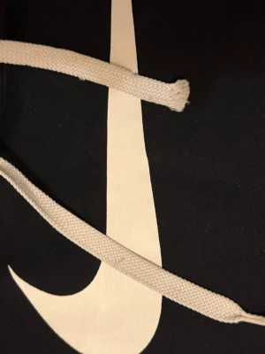 Svart hoodie med Nike-logga S - Snygg svart hoodie från Nike med stor vit Swoosh-logga på bröstet och vita dragsnören i huvan. Klassisk passform och mjukt material som känns skönt mot huden. Perfekt för dig som gillar sportig och avslappnad stil.