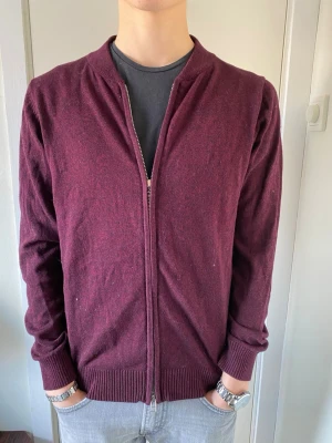 Vinröd ziptröja i ullblandning - Snygg vinröd ziptröja med ribbade muddar och krage. Tröjan har en enkel design utan synliga loggor och är tillverkad i mjuk ullblandning. Perfekt att slänga på sig över en t-shirt för en avslappnad look. Märket är från Grand Frank och storleken Small. 