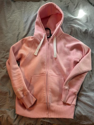 Rosa Russemerch hoodie med dragkedja - Använd 1 gång så som ny. Ljusrosa hoodie från Russemerch med dragkedja framtill, stora fickor och vit dragsko i huvan. Muddar vid ärmslut och nederkant för en skön passform. Broderad text på ena ärmen. Perfekt för en avslappnad och trendig look.