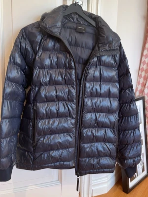 Svart dunjacka från peak performance  - Snygg svart dunjacka från peak performance  med quiltad design och hög krage. Jackan har dragkedja framtill, två sidofickor med dragkedja och en justerbar huva. Perfekt för kalla vinterdagar tack vare det varma dunfodret.