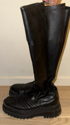 Svarta chunky knähöga boots - Säljer ett par svarta knähöga boots med chunky platåsula och grovt mönster undertill. Skorna har en slät ovandel i konstläder och coola sömdetaljer över foten. Perfekta för dig som vill ha en edgy och trendig look.