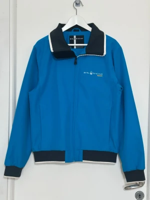 Blå vindjacka från Sail Racing - Blå vindjacka från Sail Racing med svart och vit krage, dragkedja framtill och logga på bröstet. Jackan har svarta muddar vid ärmslut och nederkant samt vita detaljer längs kragen. Tillverkad i ett lätt och vindtätt material, perfekt för blåsiga dagar.