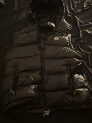 Äkta !!!! Svart dunväst från Moncler - Snygg svart dunväst från Moncler med glansig finish och klassisk logotyp på bröstet. Västen har två dragkedjefickor framtill, hög krage och huva. Perfekt för lager-på-lager och streetwear-stil. Äkta !! 