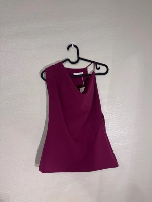  Mycket mörkrosa ärmlös topp ZARA - Säljer en stilren, mörkrosa ärmlös topp med draperad v-ringning framtill och slät baksida. Toppen har en lätt figurnära passform och är tillverkad i ett mjukt, slätt syntetmaterial. Perfekt att styla med jeans eller kjol för en trendig look.