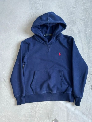 Mörkblå hoodie från Polo Ralph Lauren - Mörkblå hoodie med klassisk känguruficka och huva från Polo Ralph Lauren. Broderad röd logga på bröstet. Mjuk bomullskvalitet och ribbade muddar vid ärmslut och nederkant. Perfekt för en avslappnad och stilren look.