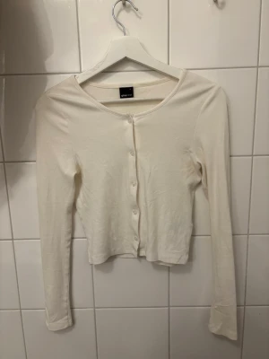 Vit kofta från Gina Tricot - Snygg och enkel vit kofta från Gina Tricot med rund halsringning och knäppning framtill. Modellen är croppad och har långa ärmar. Perfekt att styla över ett linne eller t-shirt. Mjuk och skön bomullskänsla.