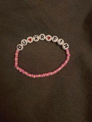 Pärlarmband med text DU ÄR FIN - Sött armband med rosa och vita pärlor, där de vita pärlorna bildar texten 'DU ÄR FIN' med röda hjärtan. Perfekt accessoar för att sprida lite extra pepp i vardagen. Elastiskt band för enkel användning.