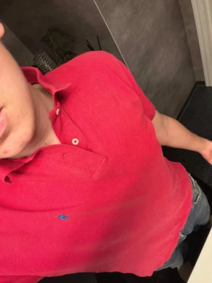 Röd pikétröja från Ralph Lauren - Klassisk röd pikétröja från Ralph Lauren med blå logga broderad på bröstet. Tröjan har krage, knappar vid halsen och korta ärmar. Perfekt till jeans för en avslappnad men snygg stil.