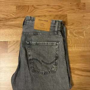 Grå raka jeans från Jack & Jones - Säljer ett par grå jeans från Jack & Jones med klassisk femficksdesign och raka ben. Jeansen har en snygg tvättad look och är tillverkade i bomull. Perfekta för en avslappnad stil.