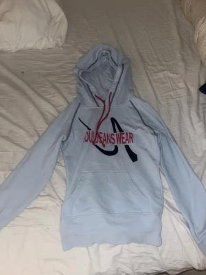 Ljusblå hoodie från Zoul - Ljusblå hoodie från Zoul med röd dragsko och tryck i rött och svart framtill där det står 'Jeans Wear'. Klassisk känguruficka och mjukt material som känns skönt mot huden. Perfekt för en avslappnad och cool stil.