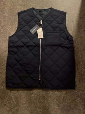 Svart quiltad dunväst  - Stilren svart dunväst från med quiltat mönster och dragkedja framtill. Västen har en bröstficka och är ärmlös med rundad hals. Perfekt lager-på-lager-plagg för kyliga dagar och enkel att matcha med olika outfits. Helt ny endast testad nypris 599