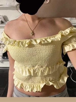 Gul offshoulder topp med volanger - Supersöt ljusgul offshoulder topp med volanger och smockad struktur. Toppen är croppad och har elastiska detaljer som ger en bekväm passform. Perfekt för sommarens alla tillfällen och ger en romantisk vibe.