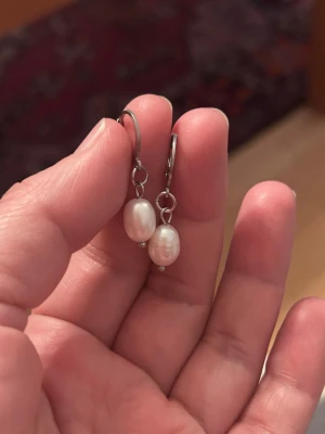 Silvriga örhängen med vita pärlor - Säljer ett par stilrena örhängen i stål silverfärg med söttvattenspärlor vita, ovala pärlor som hänger snyggt. Perfekta för dig som gillar klassiska accessoarer med en modern touch. Materialet ser ut att vara metall och pärlorna ger en elegant känsla.
