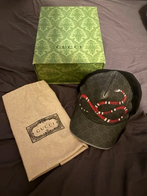 Gucci keps med orm och GG-mönster - Snygg svart keps från Gucci med klassiskt GG-mönster och en stor röd, vit och svart orm på framsidan. Kepsen har mesh på baksidan för extra ventilation och justerbar rem. Perfekt för dig som vill sticka ut med en lyxig accessoar.