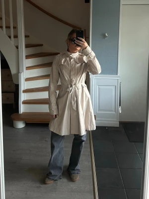 Beige trenchcoat med volangkrage - Säljer en beige trenchcoat med snygg volangdetalj vid kragen och knytband i midjan. Jackan har långa ärmar med knappar vid manschetterna och en klassisk, feminin siluett. Perfekt för dig som vill ha en trendig och stilren look.