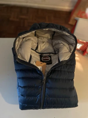 Mörkblå dunjacka från Colmar - Snygg mörkblå dunjacka från Colmar med huva och ljus insida. Jackan har dragkedja framtill, två sidofickor med dragkedja och Colmar-logga på ärmen. Perfekt för kalla vinterdagar och riktigt skön att bära.