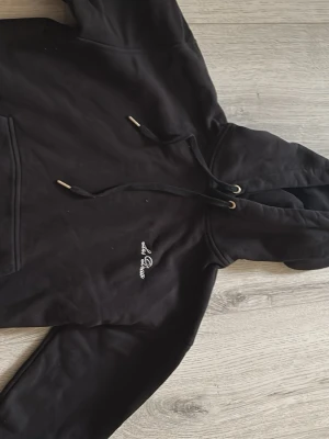 Svart hoodie LES DEUX - Svart hoodie från LES DUEX med broderad logga på bröstet. Klassisk modell med huva, dragsnören med metalländar och känguruficka framtill. Tillverkad i mjukt material, perfekt för en chill och avslappnad stil.