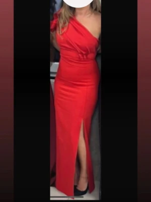 Röd oneshoulder långklänning med slits - Snygg röd långklänning med oneshoulder och markerad midja. Klänningen har en hög slits framtill och en volangdetalj vid axeln. Perfekt för dig som vill sticka ut med en elegant och modern look.