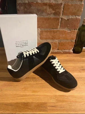 Maison Margiela svarta sneakers - INTRESSEKOLL! Svarta sneakers från Maison Margiela med klassisk design, vita snören och beige gummisula. Skorna har en mix av läder och mocka i ovandelen och en låg silhuett. Perfekta för dig som gillar stilrena och trendiga sneakers.