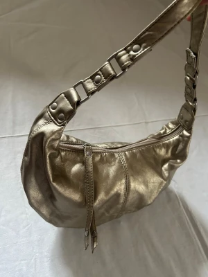 Guldig axelväska i metallic-look - Snygg axelväska i guldigt metallic-material med coola detaljer på axelremmen och dragkedja upptill. Väskan har en mjuk, rundad form och dubbla dragkedjor som ger en trendig vibe. Perfekt för dig som vill sticka ut med accessoarerna.