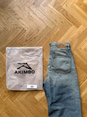Blå raka jeans från Akimbo - Säljer ett par blå raka jeans från Akimbo med klassisk femficksdesign och hög midja. Jeansen har en avslappnad passform och är tillverkade i denim med snygg tvättad look. Perfekta för en trendig och avslappnad stil.