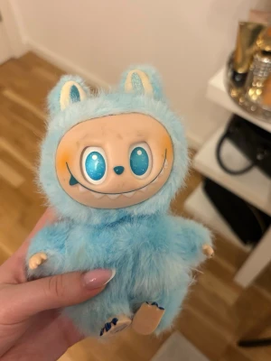 Blå Labubu  - Söt och unik monsterfigur med blått fluffigt pälsmaterial, stora glittriga blå ögon och ett brett leende. Figuren har små öron och tassar, samt en beige ansikte med tecknade detaljer. Perfekt som prydnad eller samlarobjekt.