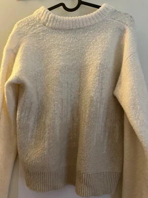Beige stickad tröja med rund hals - Mysig stickad tröja i beige med rund halsringning och ribbstickade muddar vid ärmslut och nederkant. Tröjan har en avslappnad passform och är perfekt för lager-på-lager. Materialet känns mjukt och fluffigt, vilket gör den extra skön att bära.