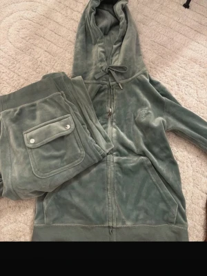 Grågrön velour loungewear-set Juicy Couture - Mysigt loungewear-set i grågrön velour från Juicy Couture. Setet består av en ziphoodie med dragsko i huvan och matchande byxor med fickor och silverfärgade knappar. Båda delarna har broderad JC-logga framtill. Perfekt för chill dagar hemma. Storlek XS på både, äkta sett 