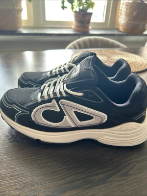 Dior B30 svarta sneakers - Snygga Dior B30 sneakers i svart med vita detaljer och vita snören. Skorna har en sportig design med mesh och läderpaneler samt tydlig Dior-logga på sidan. Tillverkade i Italien och perfekta för dig som gillar exklusiva sneakers.