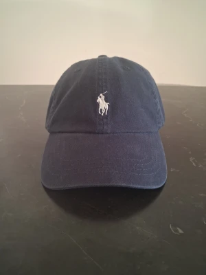 Mörkblå keps från Polo Ralph Lauren - Snygg mörkblå keps från Polo Ralph Lauren med klassisk vit broderad logga framtill och 'POLO' broderat bak. Justerbar rem baktill för perfekt passform. Tillverkad i bomull för en bekväm känsla.