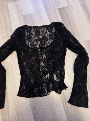 Svart spetsblus med knytning - Trendig svart långärmad topp i spets med blommigt mönster och knytning framtill. Figurnära passform och lätt transparent material, perfekt för att skapa en edgy look.