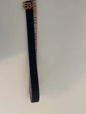 Burberry vändbart bälte med monogram - Snyggt vändbart bälte från Burberry med klassiskt rutigt mönster på ena sidan och svart monogram på andra. Spännet är i guld med Burberrys ikoniska logga. Perfekt accessoar för att lyfta din outfit.