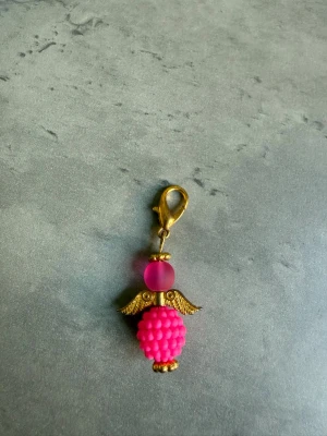 Rosa ängelberlock med guldfärgade vingar - Söt berlock formad som en ängel med guldfärgade vingar och detaljer. Kroppen består av en klotrund, bubblig pärla i starkt rosa och huvudet av en matt rosa pärla. Perfekt att fästa på väska, nyckelring eller smycke. Handgjord av mig!