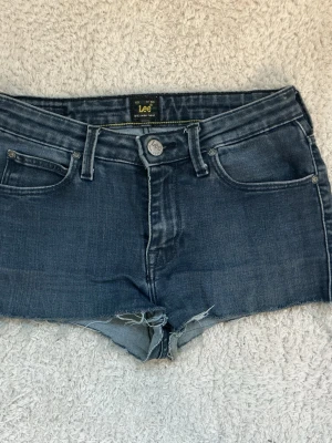 Blå jeansshorts från Lee - Säljer ett par klassiska blå jeansshorts från Lee med rå kant nertill och femficksmodell. Shortsen har knappgylf och bälteshällor, samt Lee-logga på bakfickan. Perfekta för varma dagar och enkel att matcha. Lågmidjade.
