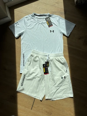 Vitt träningsset från Under Armour - Snyggt vitt träningsset från Under Armour med t-shirt och shorts. Setet har tryckta loggor och detaljer i svart och holografiskt, samt elastisk midja på shortsen. Perfekt för gym eller löpning. Materialet är lätt och andas, vilket gör det bekvämt under träning.