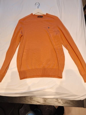 Orange stickad tröja från Gant - Snygg orange stickad tröja från Gant med rund halsringning och diskret logga broderad på bröstet. Tröjan har långa ärmar och ribbade muddar vid ärmslut och nederkant. Perfekt för dig som gillar stilrena och färgglada plagg.