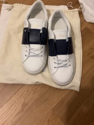 Valentino vita sneakers med blå detalj - Snygga vita sneakers från Valentino med en bred mörkblå läderdetalj över snörningen. Skorna har klassisk låg profil, vita skosnören och diskret logga på plösen. Tillverkade i slätt läder för en clean och exklusiv look. Endast skorna tillkommer 
