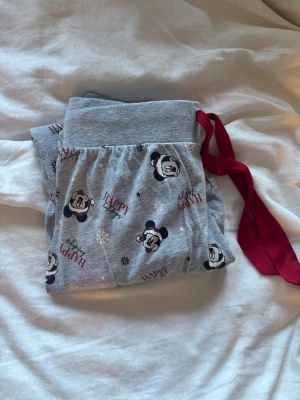 Grå Disney pyjamasbyxor med Musse Pigg - Mysiga grå pyjamasbyxor med Musse Pigg-motiv och texten 'HAPPY' i rött och svart. Byxorna har bred mudd i midjan och en röd knytband för extra komfort. Perfekta för chillkvällar hemma och har ett vinterinspirerat mönster med snöflingor.