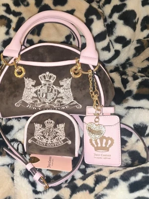 Juicy Couture brun och rosa handväska - Superfin handväska från Juicy Couture i brun mocka med ljusrosa detaljer och guldfärgade metalldelar. Väskan har broderad logga, matchande plånbok och korthållare med glittriga detaljer. Perfekt accessoar för dig som gillar lyxig och trendig stil.