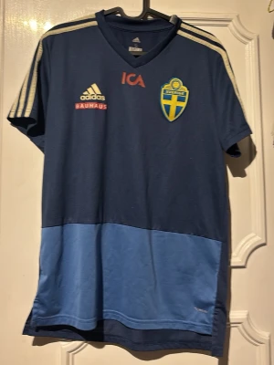 Blå Sverige Adidas träningströja - Mörkblå och ljusblå träningströja från Adidas med Sveriges landslagsemblem på bröstet. Tröjan har V-ringning, korta ärmar och klassiska Adidas-ränder på axlarna. Tryck med ICA och Bauhaus på framsidan. Perfekt för fotbollsträning.